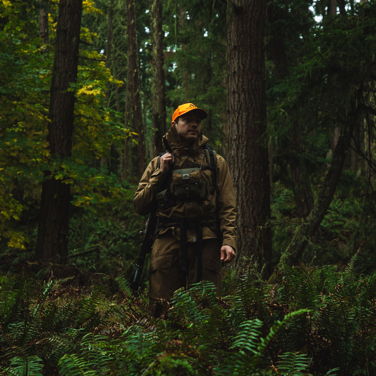 A man wears a drab green hooded outdoor jacket, matching pants, a dark multi-pouch chest rig, and an orange cap in a forest.