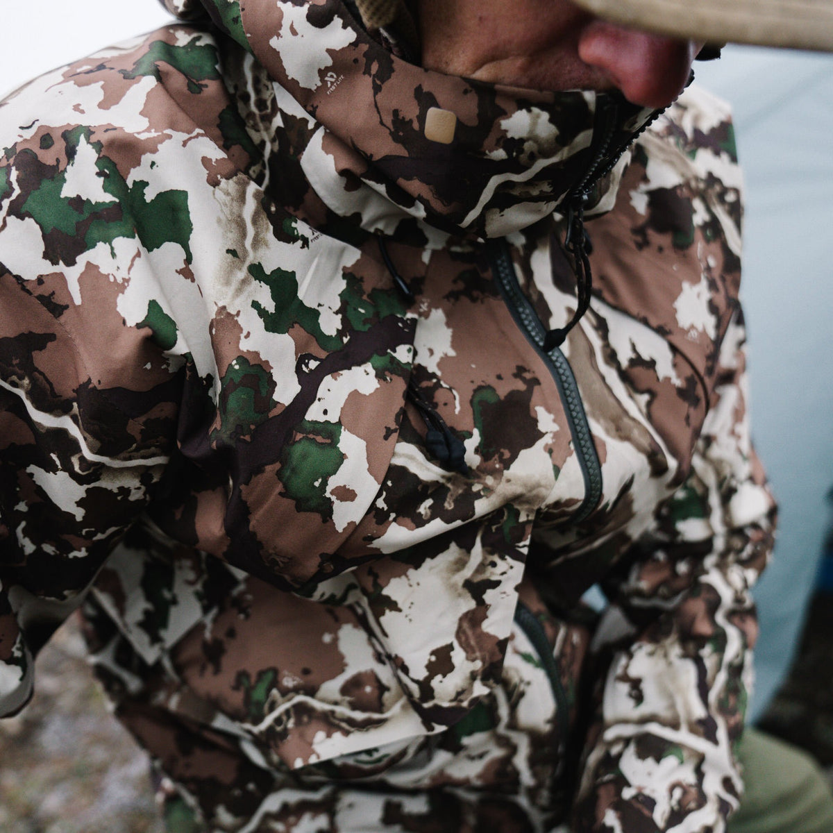 White, brown, and green camouflage hooded jacket with exposed zippers and a small square neck patch.