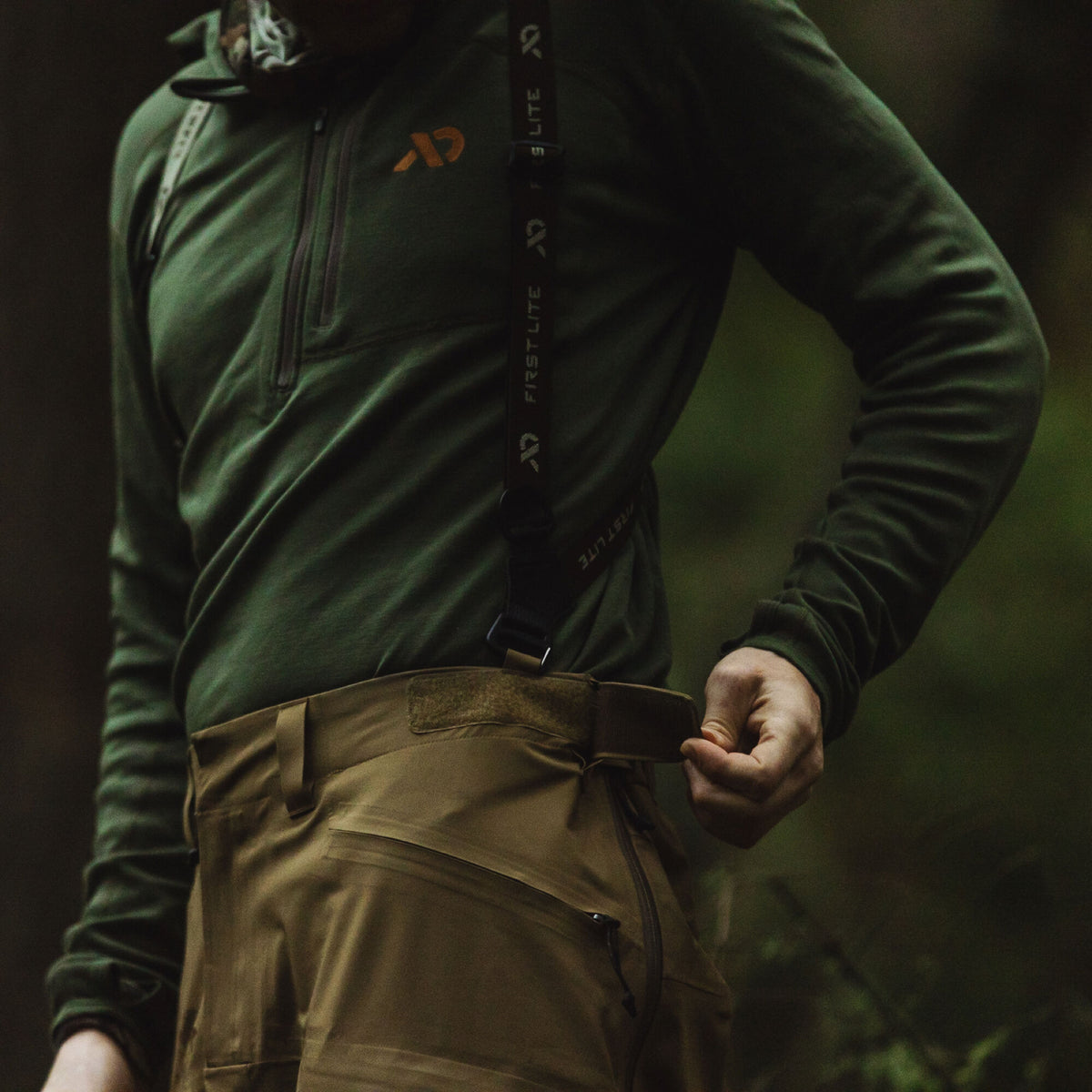 Dark green quarter-zip top, tan pants, and black First Lite suspenders on a person adjusting the pants' waist.