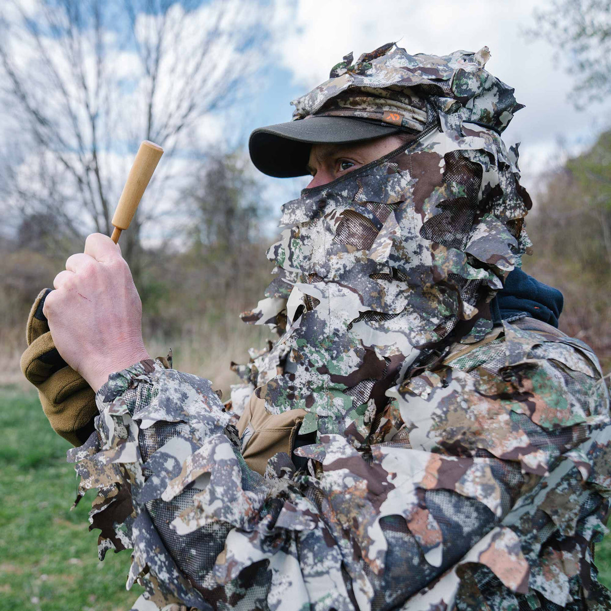 Person in a multi-color leafy 3D camouflage ghillie suit holding a wooden game call.