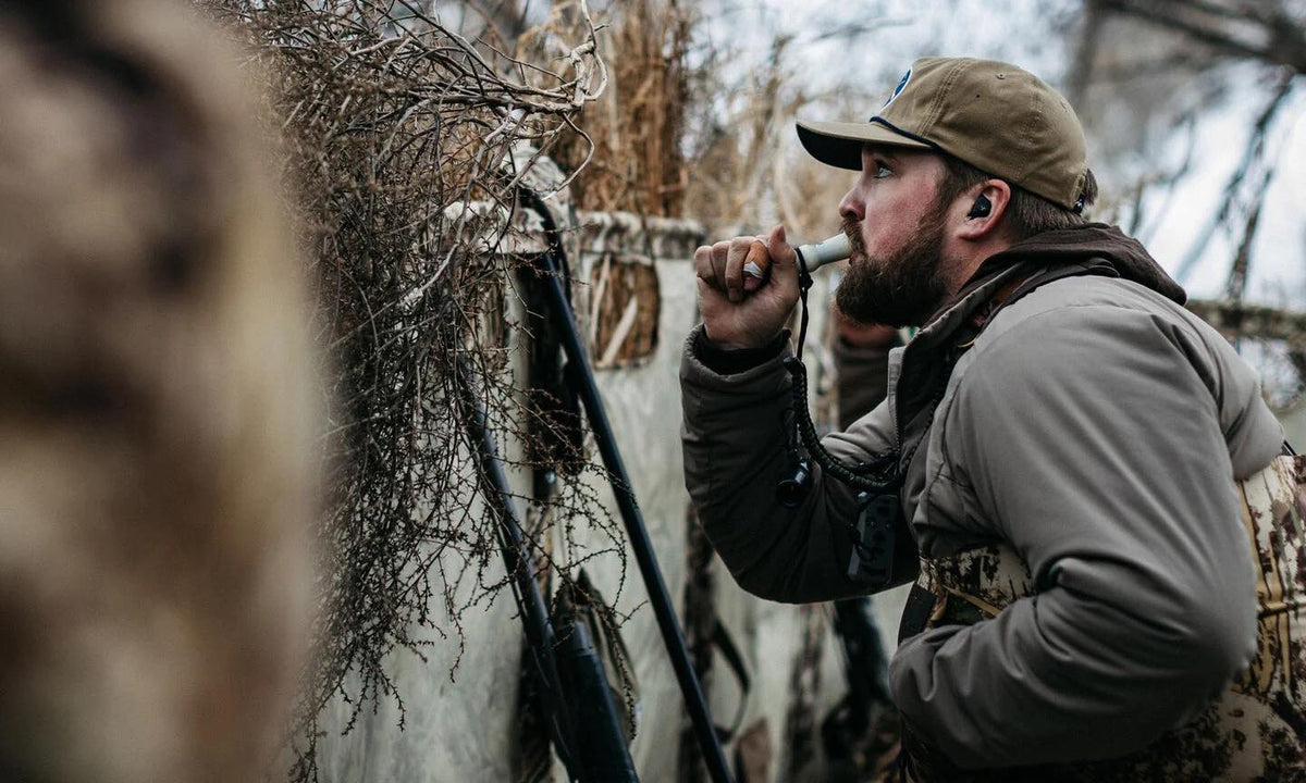 Bearded hunter uses black in-ear hearing protection with a white duck call.