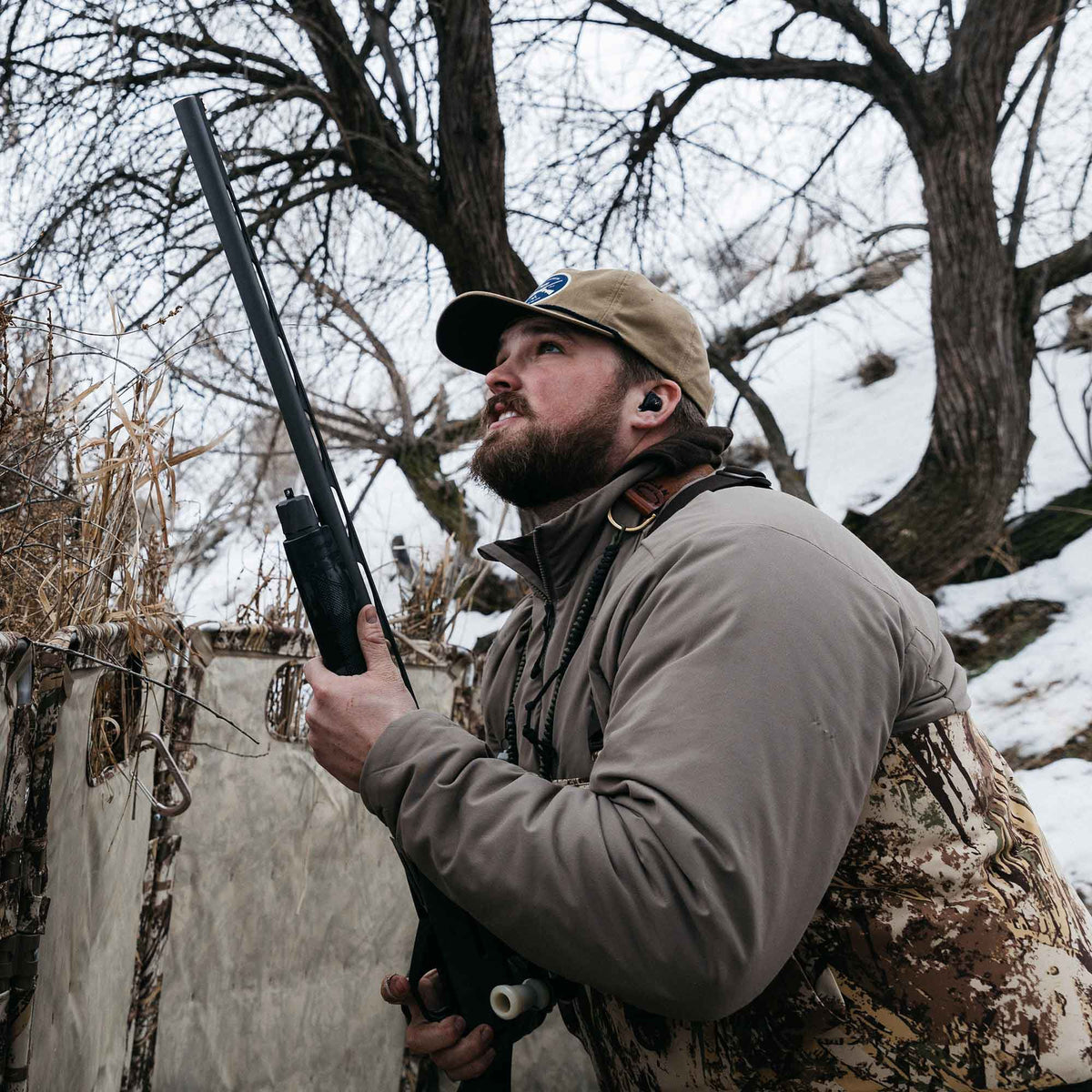 A hunter wears black in-ear hearing protection earbuds. With a walnut vest and marsh camo waders. 