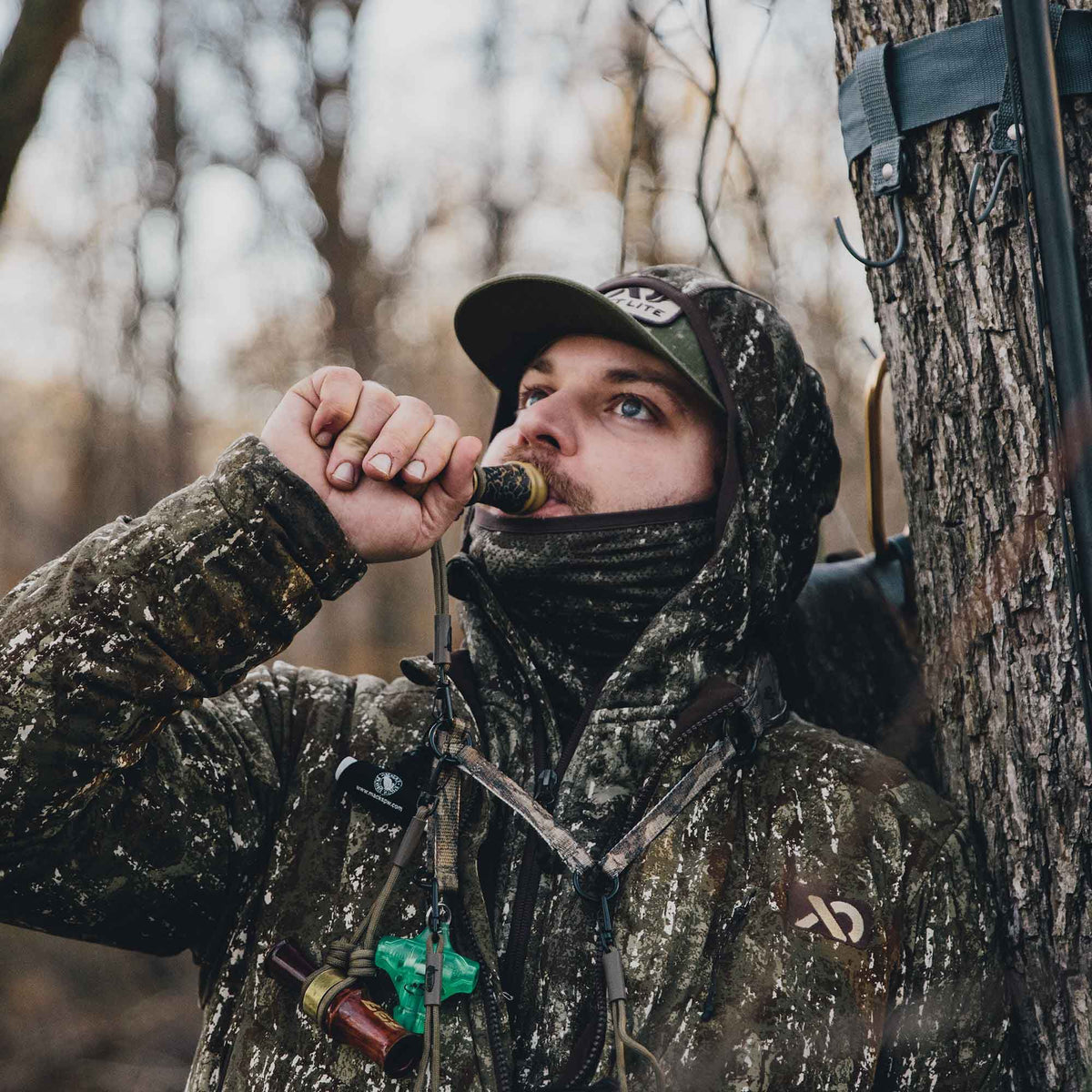 Hunter in a green and brown camouflage hooded jacket and cap blowing a duck call, with other calls hanging around his neck.