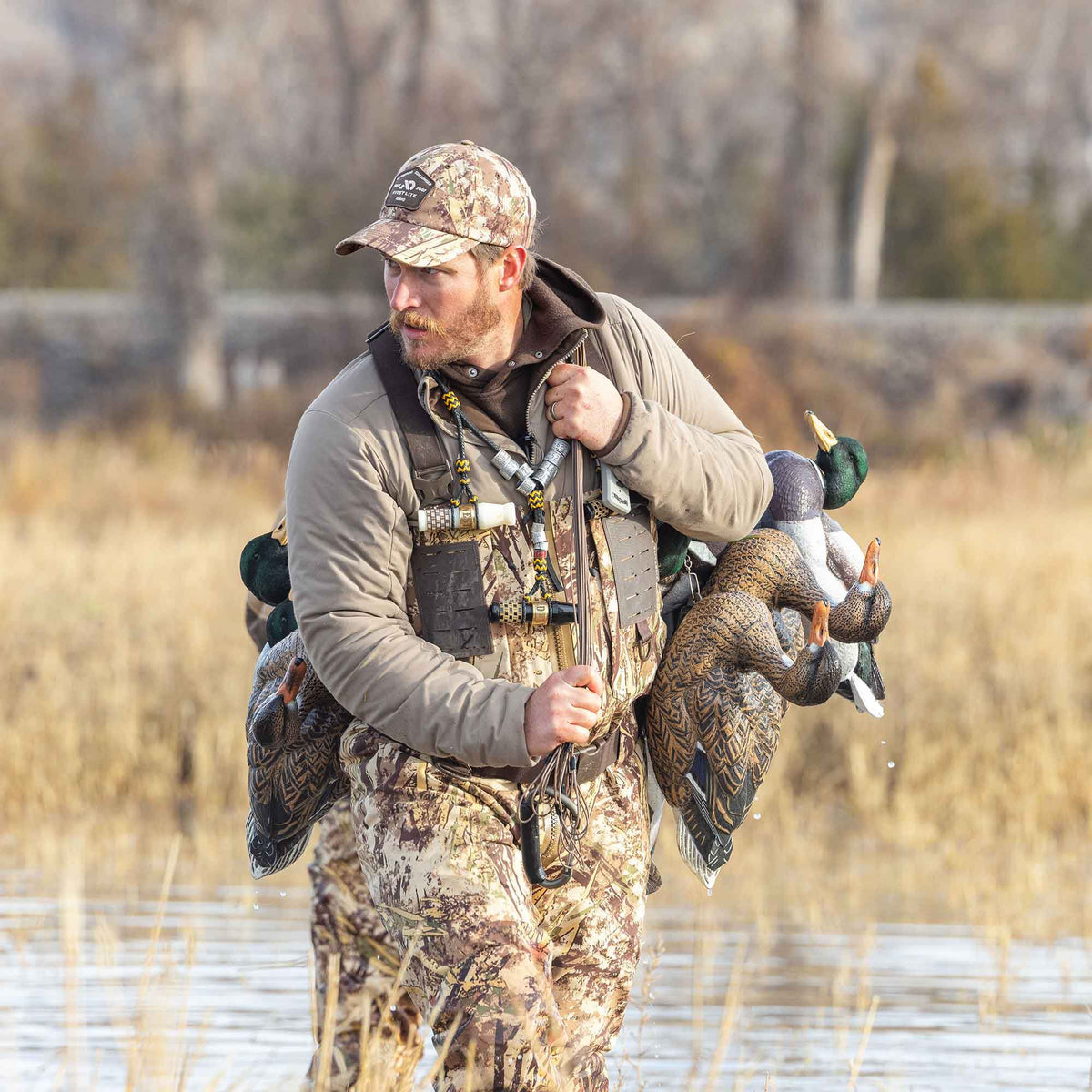 A hunter in a tan jacket, First Lite camouflage waders and hat carries several realistic duck decoys and wears a lanyard with multiple duck calls.