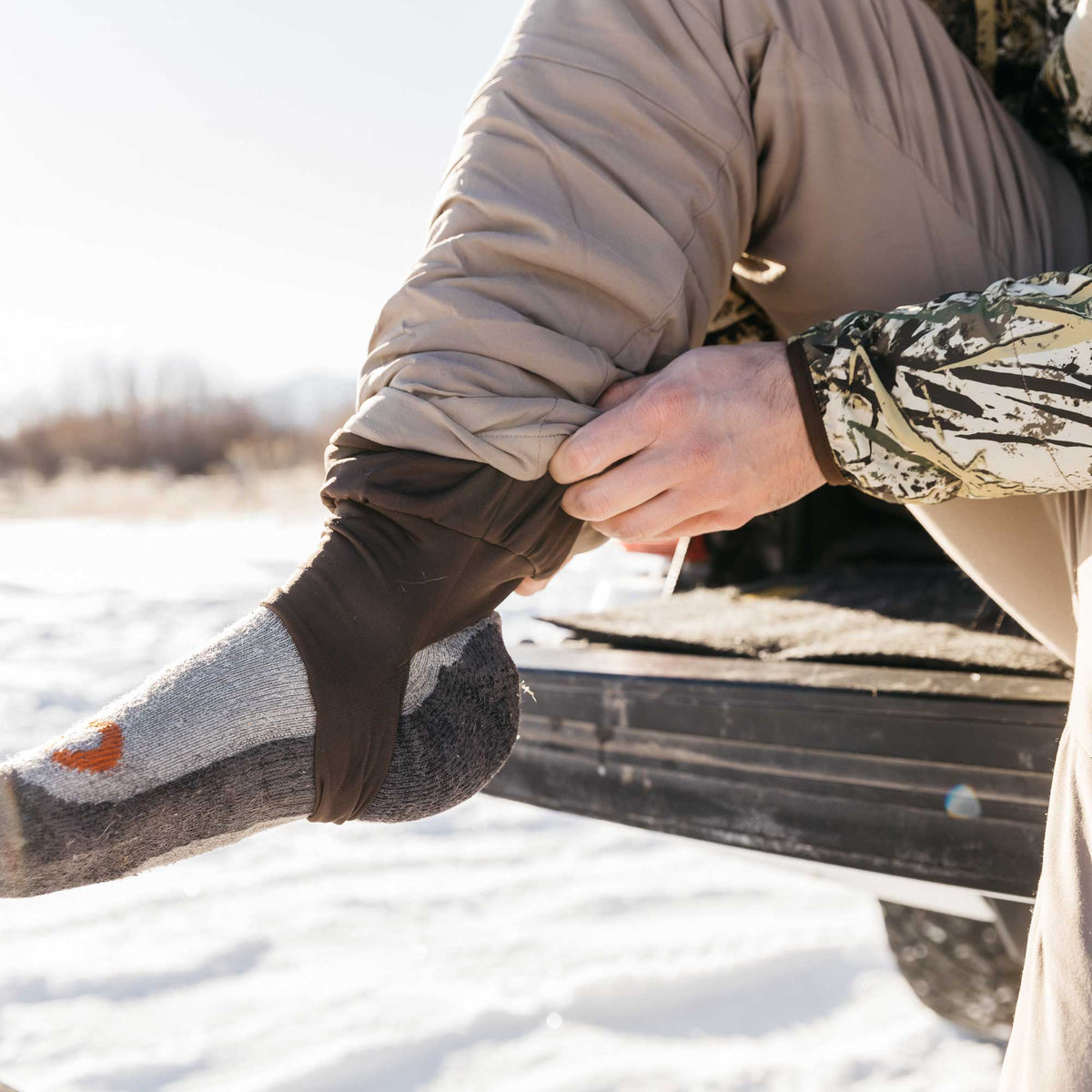 A person pulls a dark brown sock liner up their leg over a grey and dark grey sock with an orange toe logo, in a snowy environment.