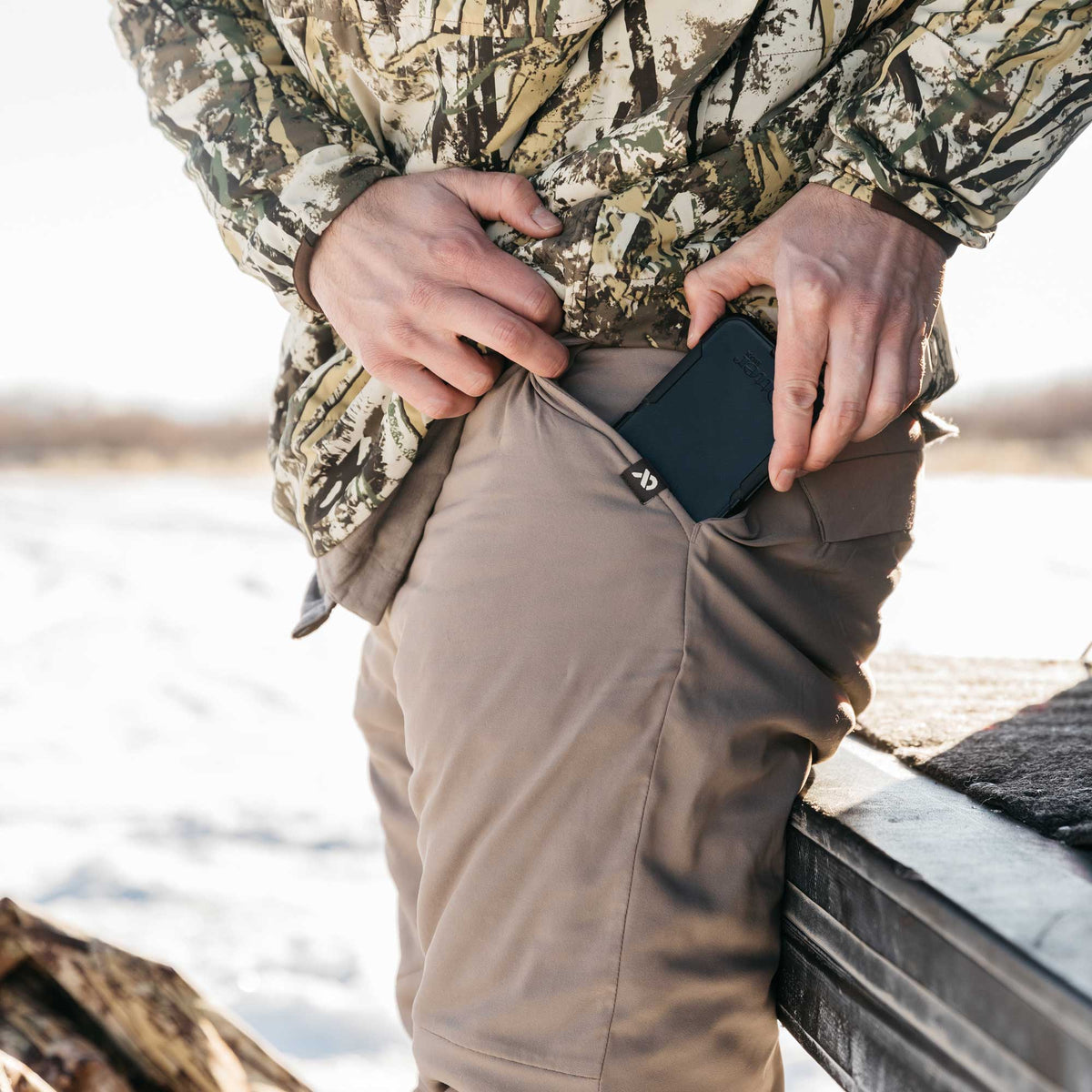 Tan outdoor pants with a phone being inserted into a zippered side pocket that has a black logo tag.