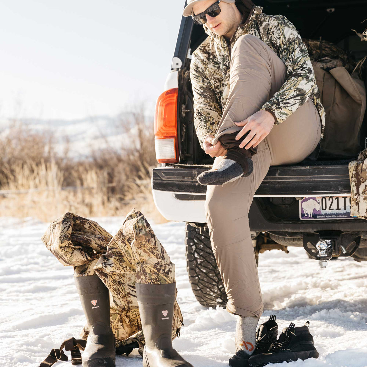 A man in a camouflage jacket and tan pants sits on a truck tailgate, adjusting his lower leg, while dark olive green Sno Goose rubber boots with a pink heart logo and black hiking boots rest on the snow.