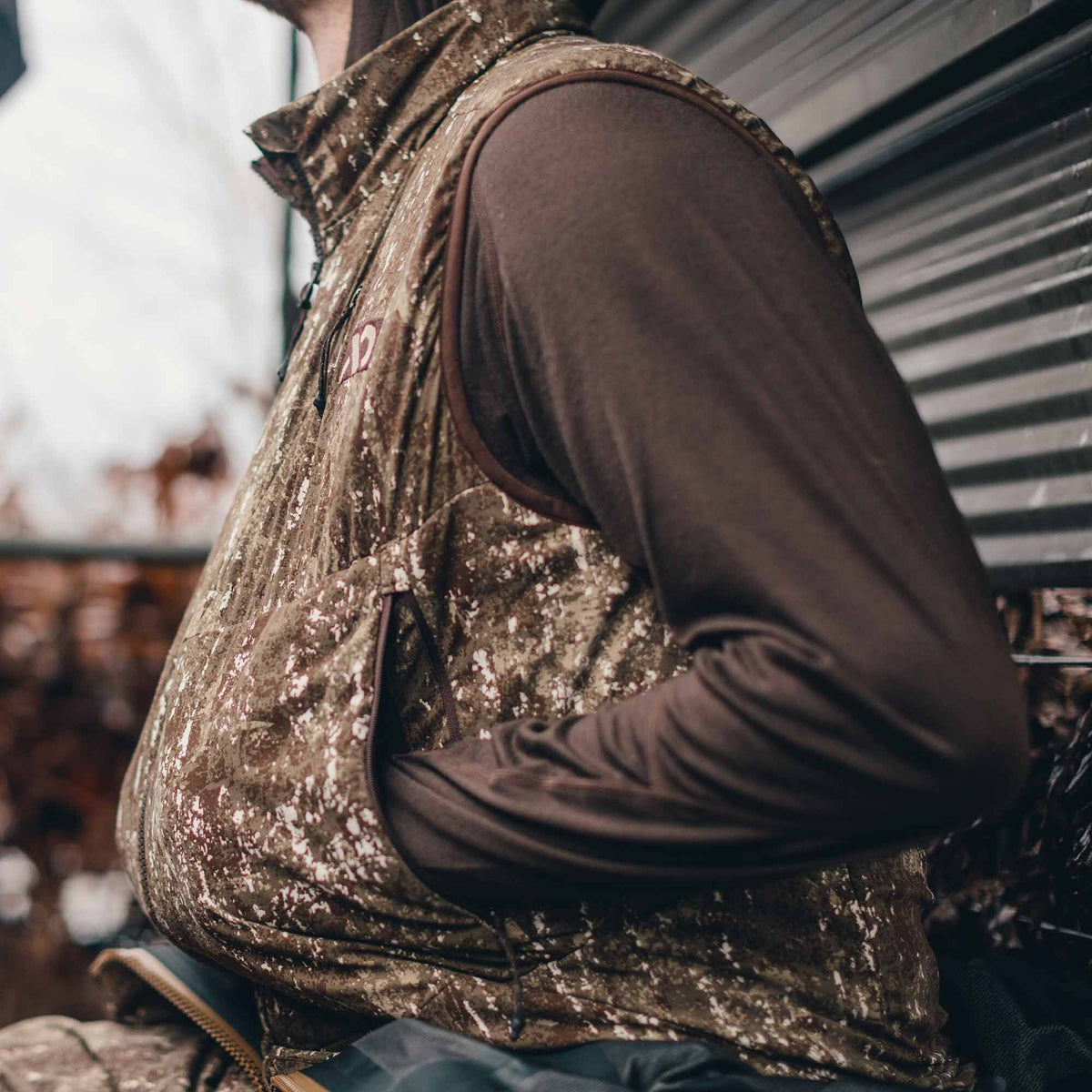 A person wears a brown and white camouflage vest with zippered pockets and a small red chest logo over a brown long-sleeve shirt.