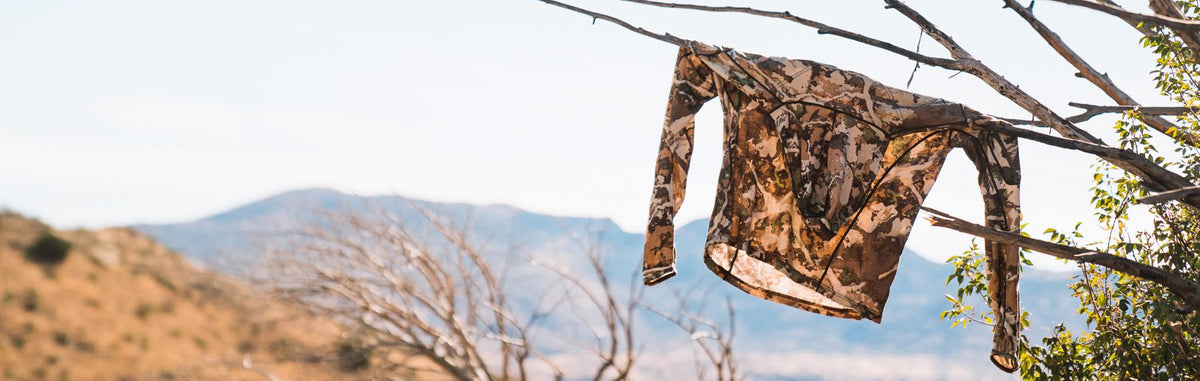 Hunting camouflage long-sleeve shirt, earth-toned with black trim, hanging on a branch.