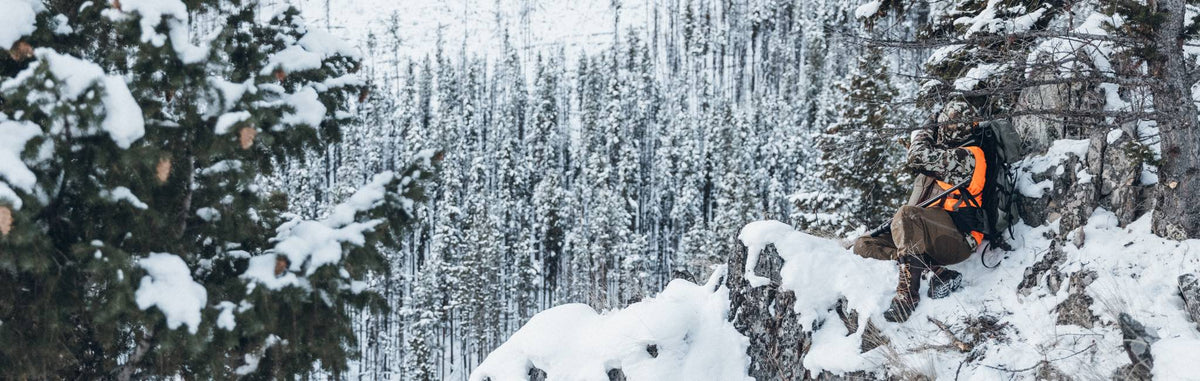 Camo hunting backpack with bright orange safety panel worn by hunter in snowy woods.