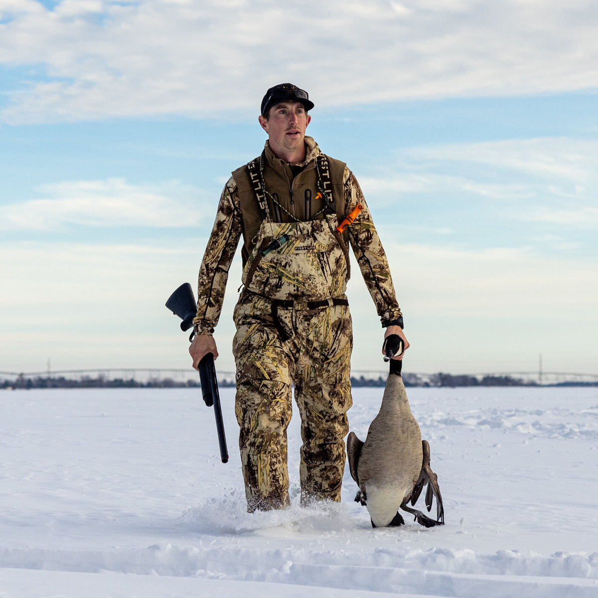 A hunter in First Lite camouflage hunting bibs with branded suspenders and a matching jacket walks through snow, holding a shotgun and a harvested goose.