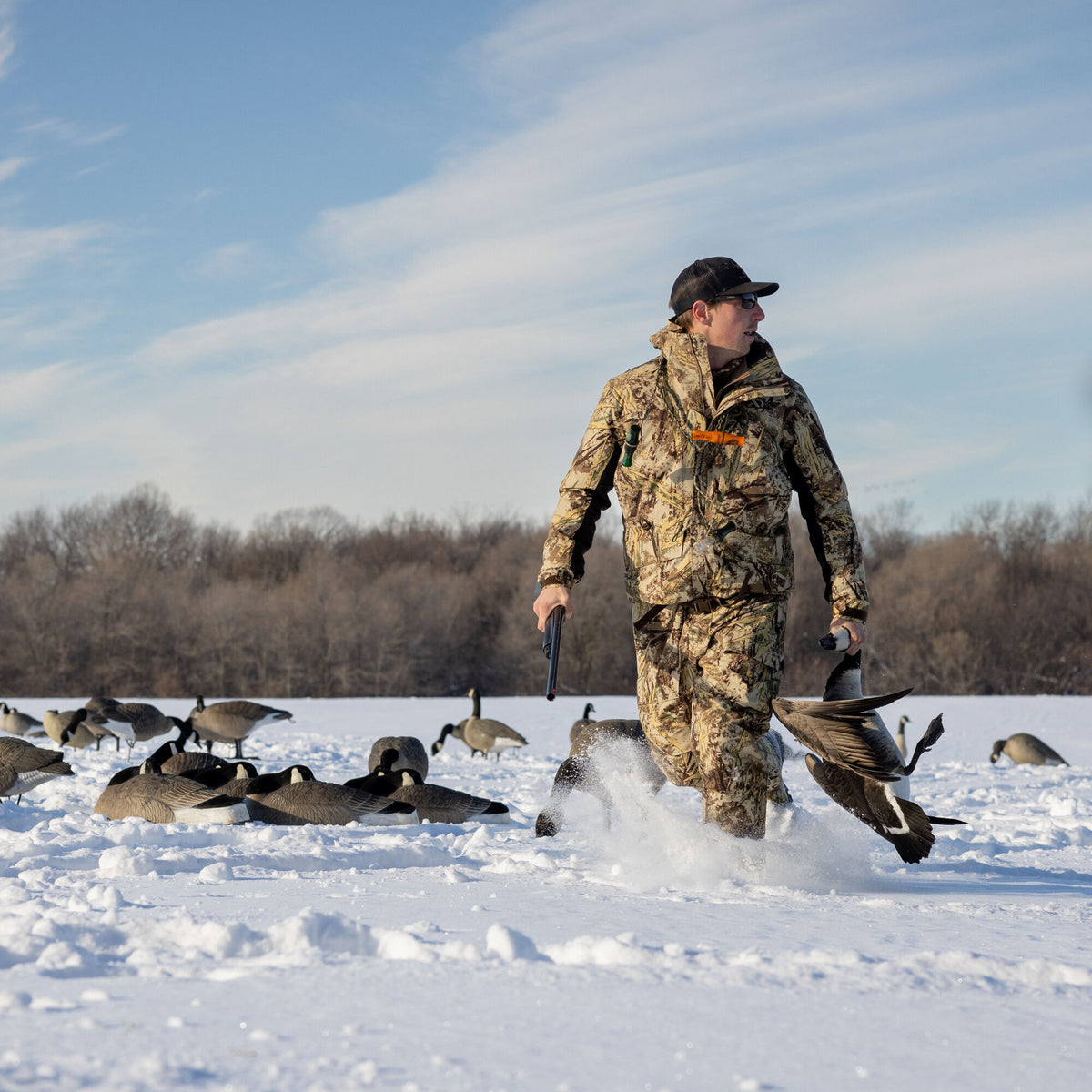 Natural camouflage hunting jacket and pants worn by a hunter walking through snow, holding a shotgun and a harvested goose.