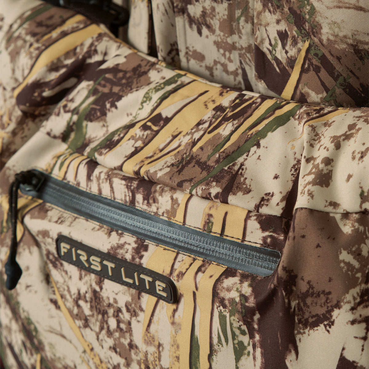 First Lite hunting apparel in brown, green, and tan camouflage, featuring a zippered pocket and the "FIRST LITE" brand logo.