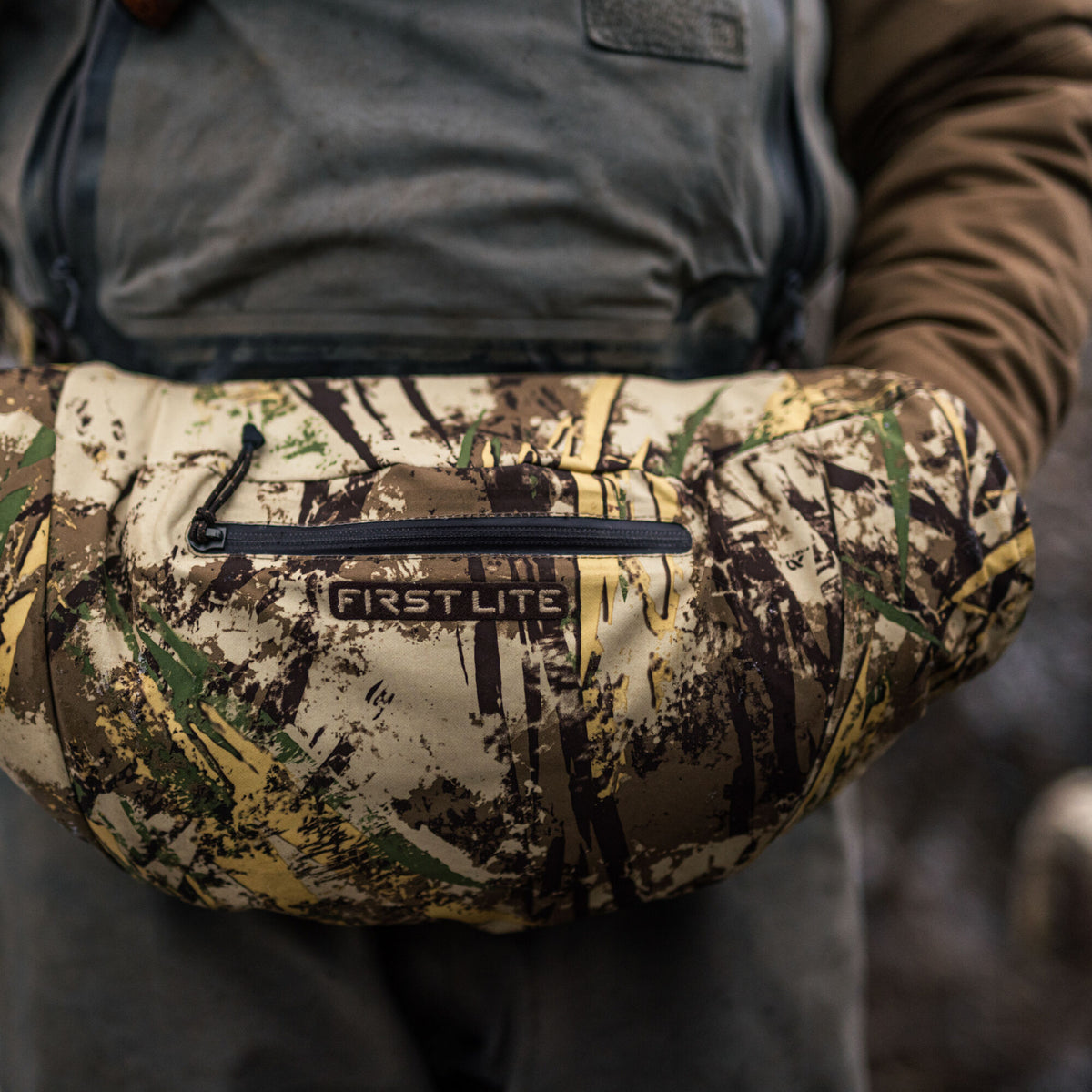 First Lite Typha camouflage hand muff with a black zippered pocket and logo, held by a person.