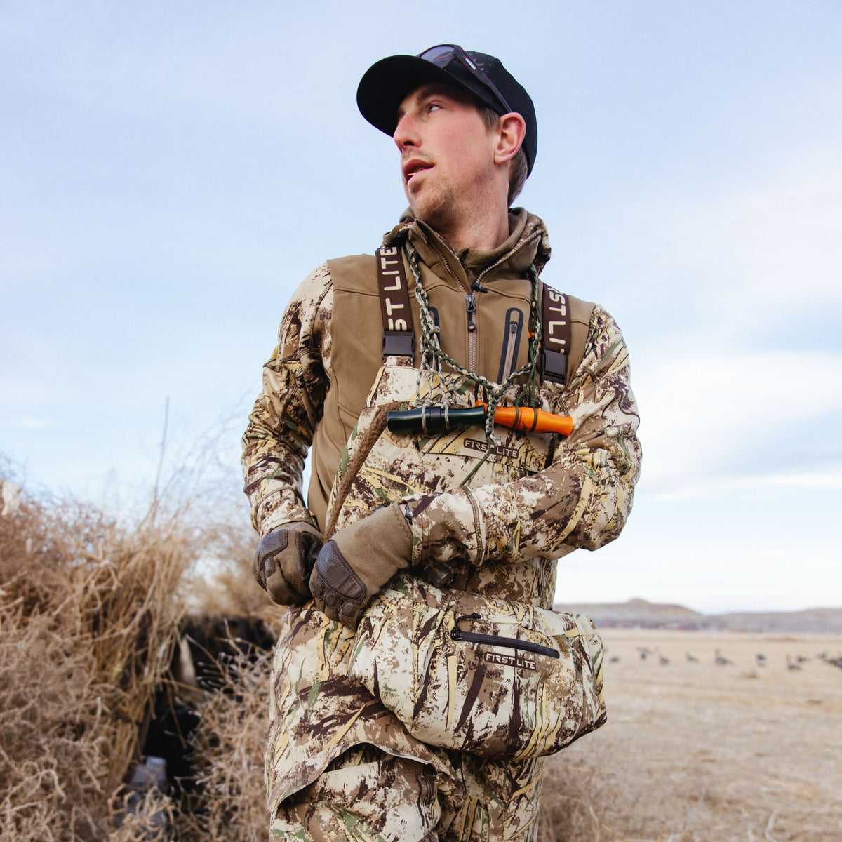 Man in First Lite Typha camouflage hunting waders, shirt, and hand warmer muff, with a brown vest and duck calls on a lanyard.