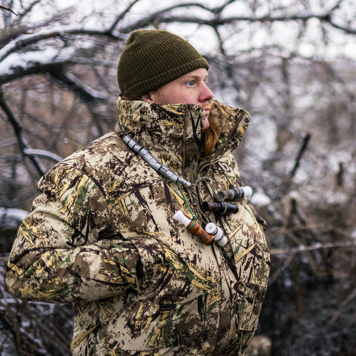 A person wears a tan and green camouflage hunting jacket with a high collar, featuring duck calls and bird bands around their neck.