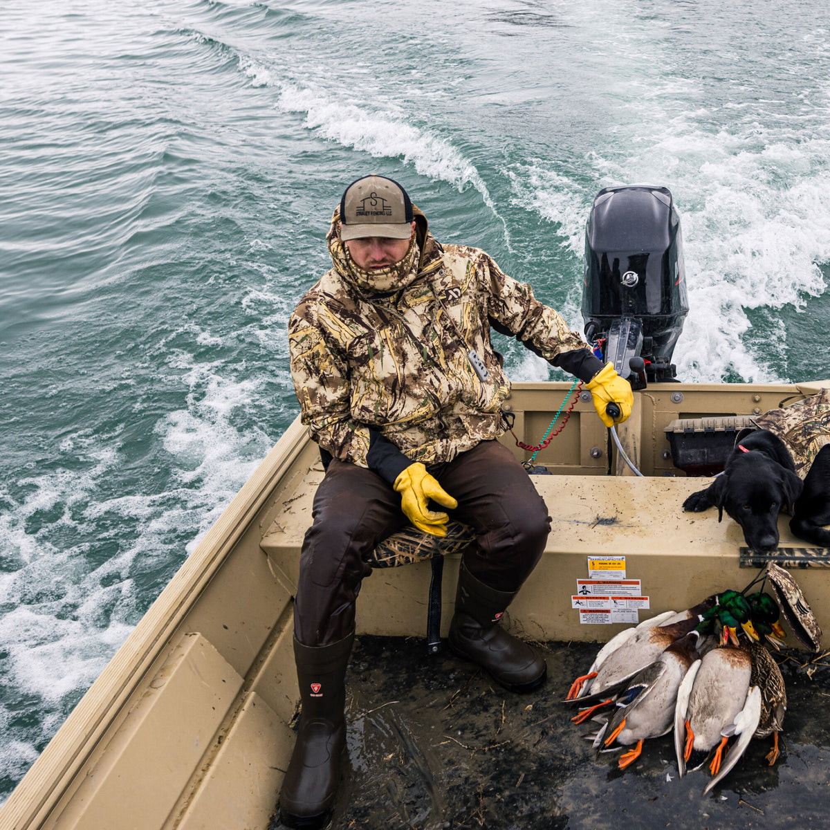 A hunter in a tan camouflage jacket, dark brown pants, and dark rubber boots sits in a tan boat with harvested ducks and a black Labrador retriever.