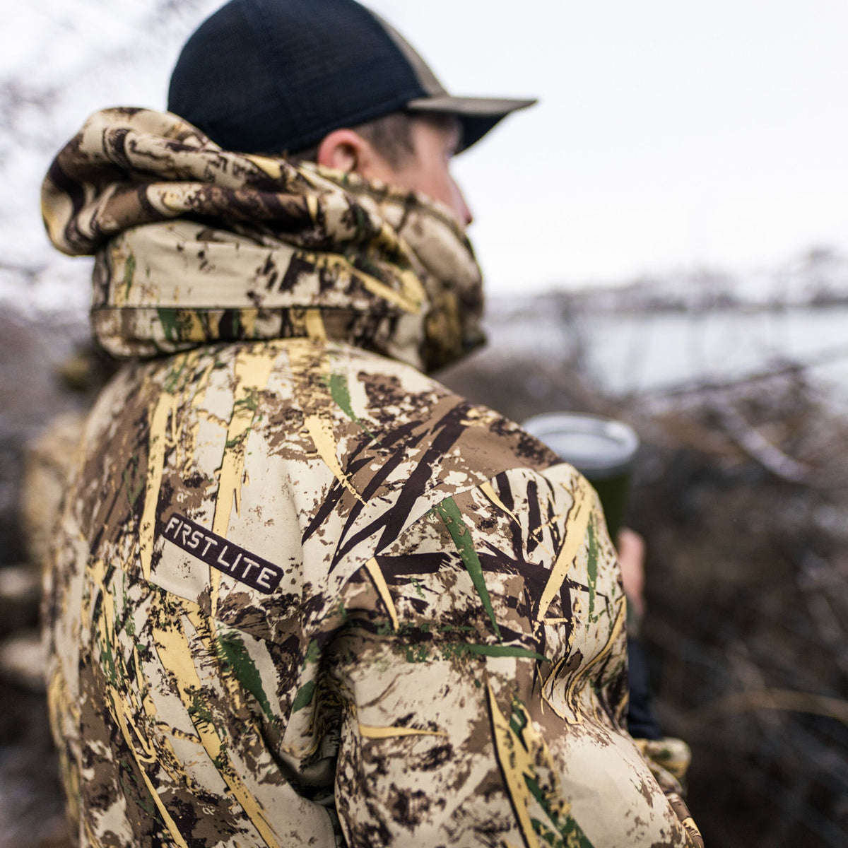 Back of a person wearing a hooded First Lite camouflage jacket with tan, brown, black, and green patterns.