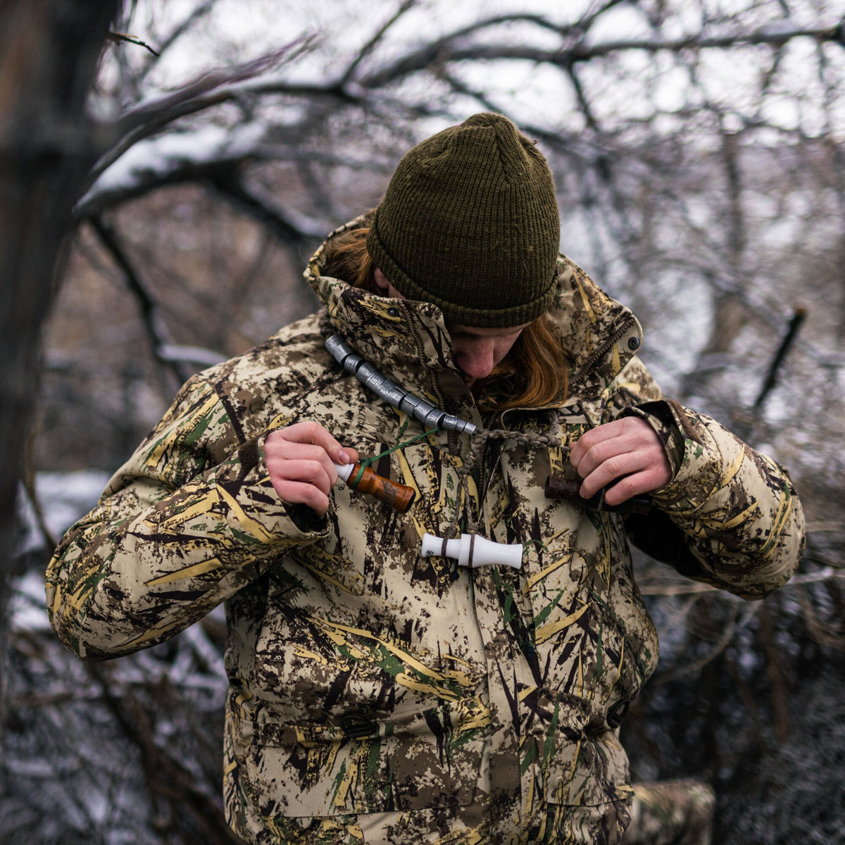 A person in an olive green beanie and a tan camouflage hunting jacket holds duck calls and metal bands on a lanyard.