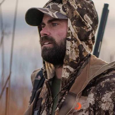 Camo hooded jacket on bearded man wearing a cap, with rifle on shoulder.