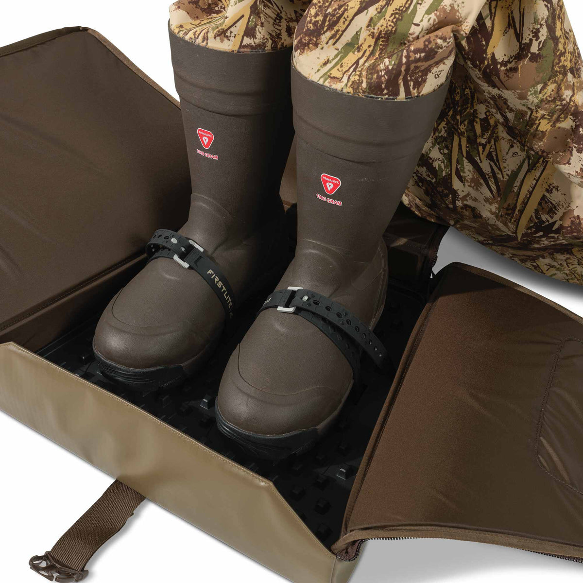A pair of dark brown PRIMALOFT 1000 GRAM insulated boots are secured with black FIRSTLITE branded straps inside an open tan and brown soft-sided boot case.