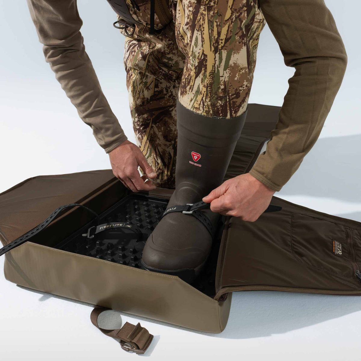 A person adjusts a strap on a dark brown First Lite wader boot featuring PrimaLoft 1000 gram insulation, worn with camouflage waders.