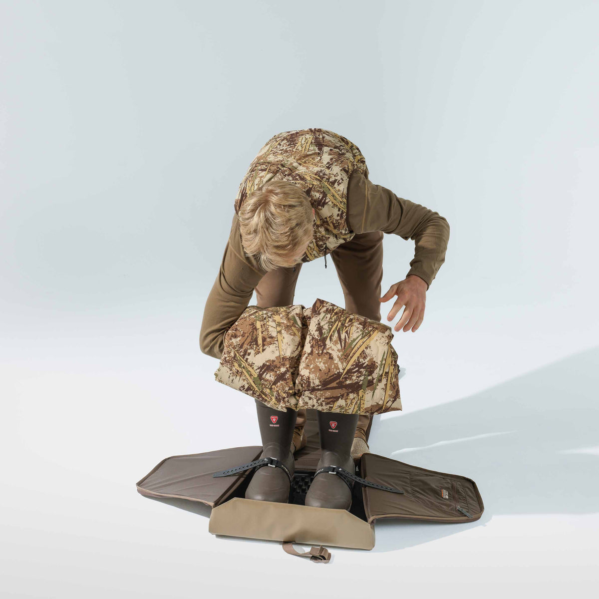 Walnut foldable boot bag and changing mat, holding two brown boots with black straps, used by a person wearing camouflage.