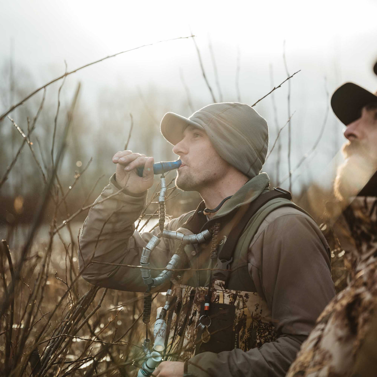 A man in hunting gear uses a blue duck call, connected to a lanyard adorned with metal bands, amidst bare branches outdoors.