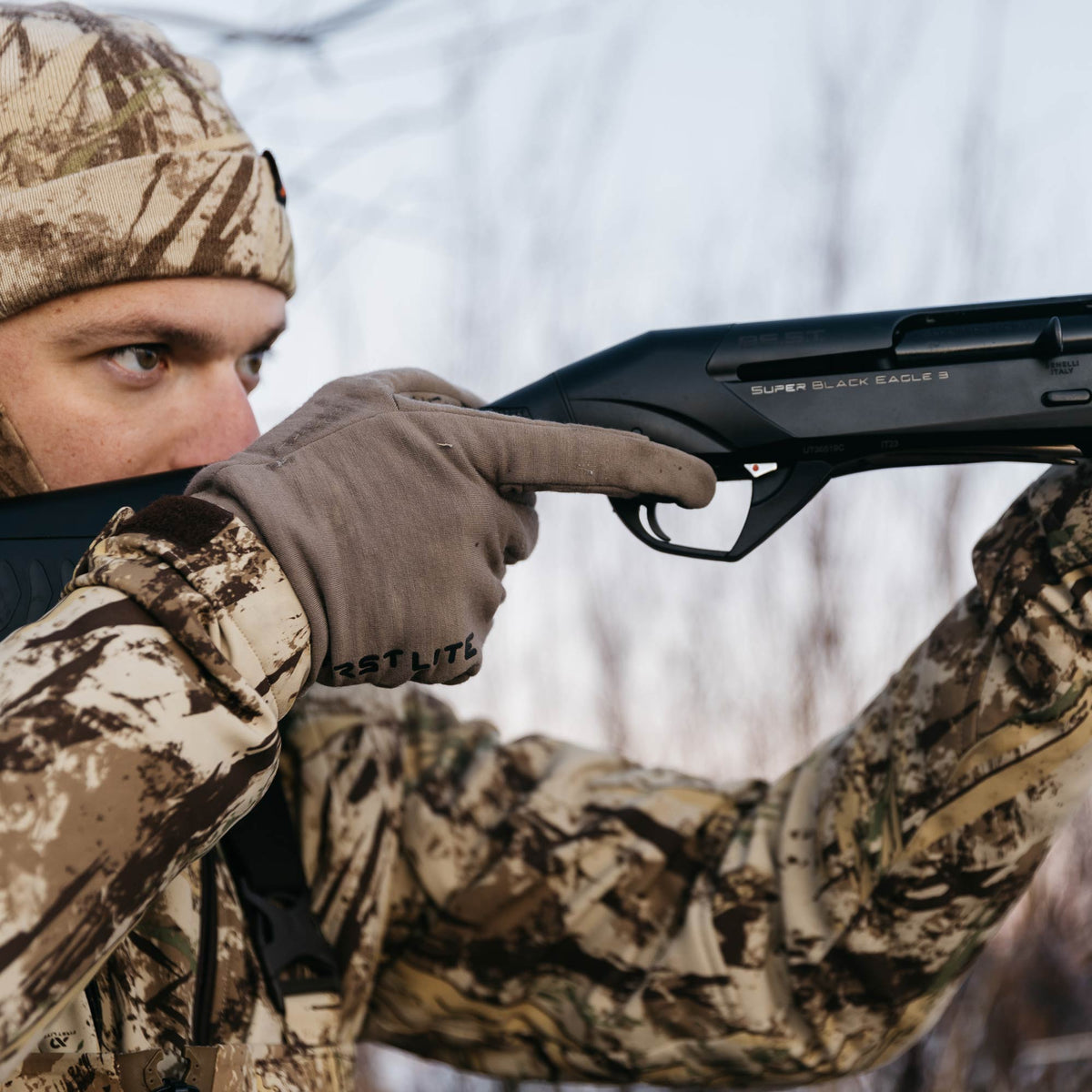 A person aims a black Benelli Super Black Eagle 3 shotgun with a visible red safety button near the trigger.