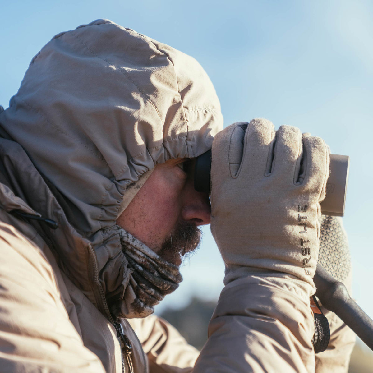 Tan binoculars held by a person wearing tan gloves with "FIRST LITE" text and a hooded jacket, against a clear sky.