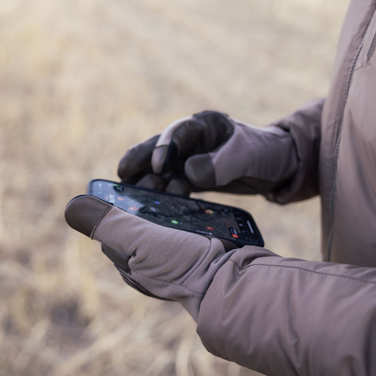 A close-up of a person wearing gray tan and leather touchscreen gloves, holding a smartphone outdoors. 