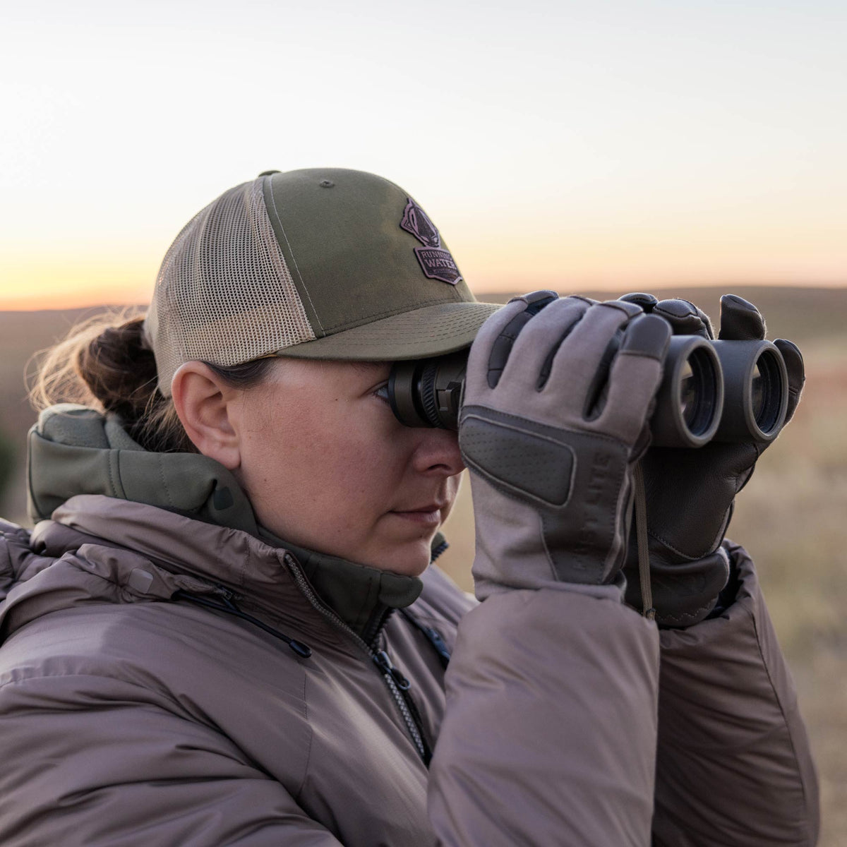 Image showing a woman looking through a pair of binoculars in a puffy jacket, hat, and hybrid gloves. 