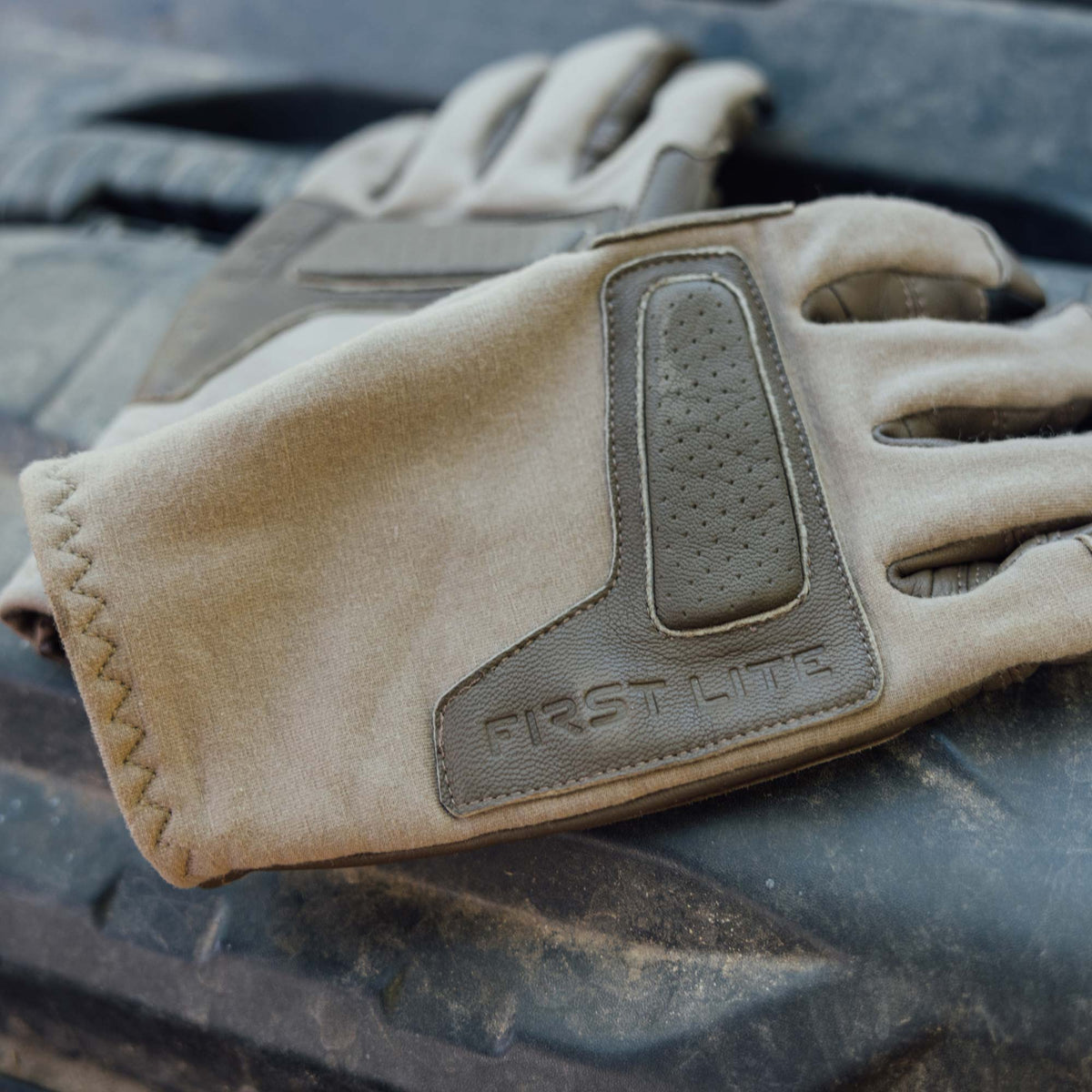 A close-up of a tan leather tactical glove with leather protective panels, including a textured grip panel and an embossed 'FIRST LITE' logo, resting on a black rubber tire.