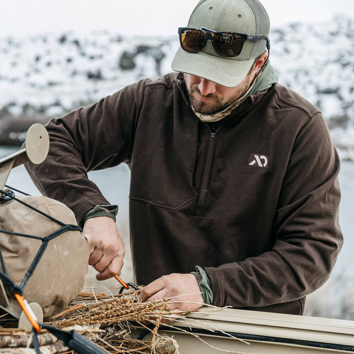 Dark brown quarter-zip pullover with a subtle diamond texture and white logo on the left chest, worn by a man preparing outdoor gear.