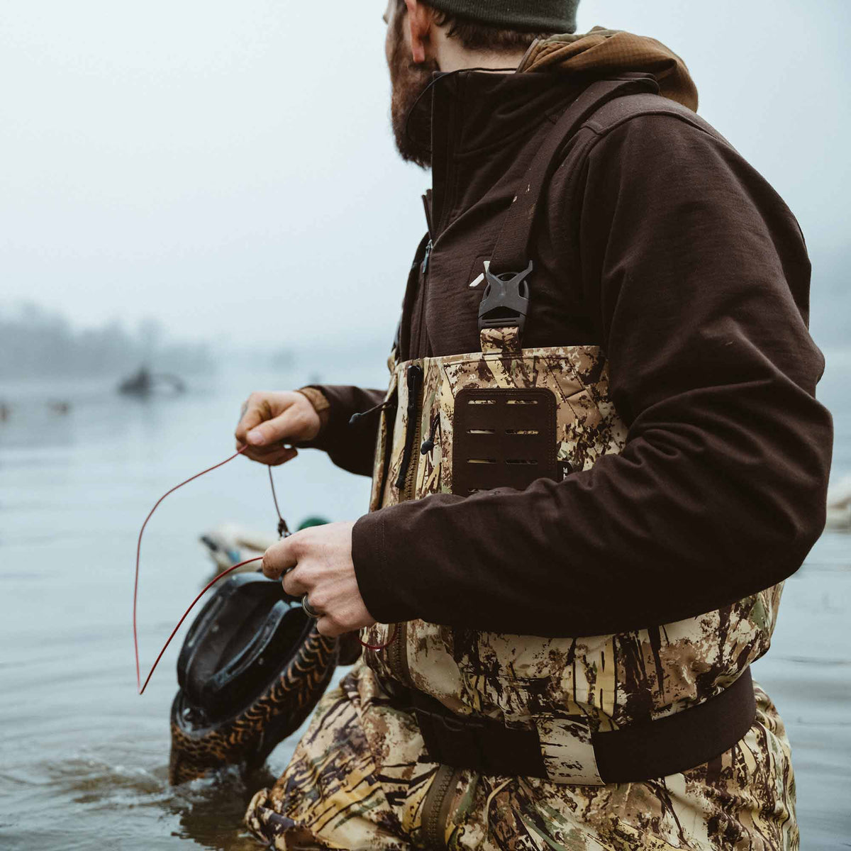 A person in camouflage waders featuring a chest attachment panel and a brown hooded jacket stands in water holding a dark decoy.