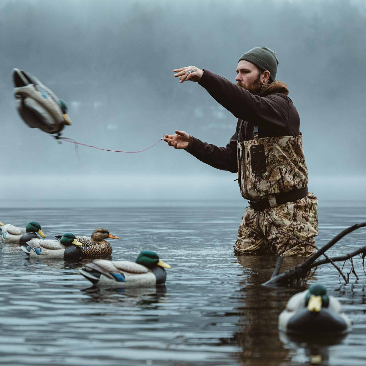 Man wearing earthy camouflage chest waders with a black chest pouch, deploying duck decoys in water.