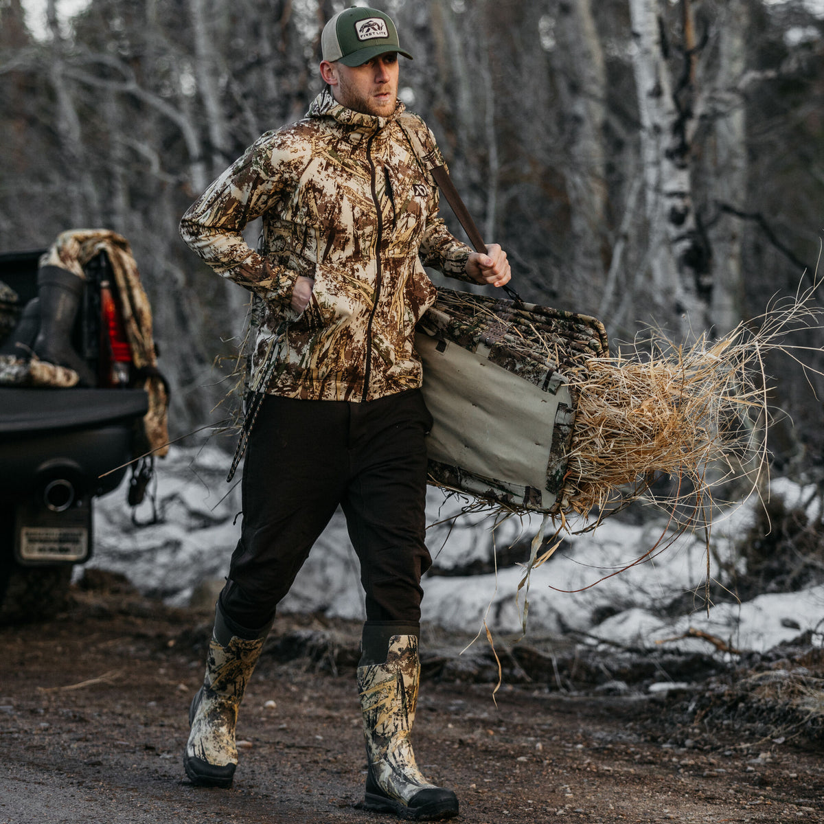 A man wears a brown and tan camouflage hooded jacket, dark pants, and tall matching boots while carrying a large, grass-covered camouflage hunting blind over his shoulder.