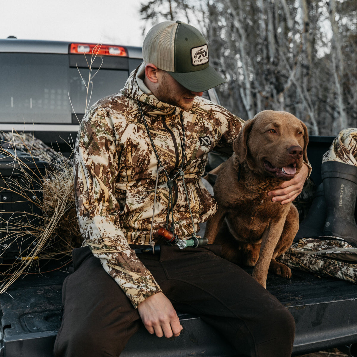 Men's full-zip hunting hoodie in brown, tan, and green camouflage, worn by a man sitting with a brown dog and duck calls.