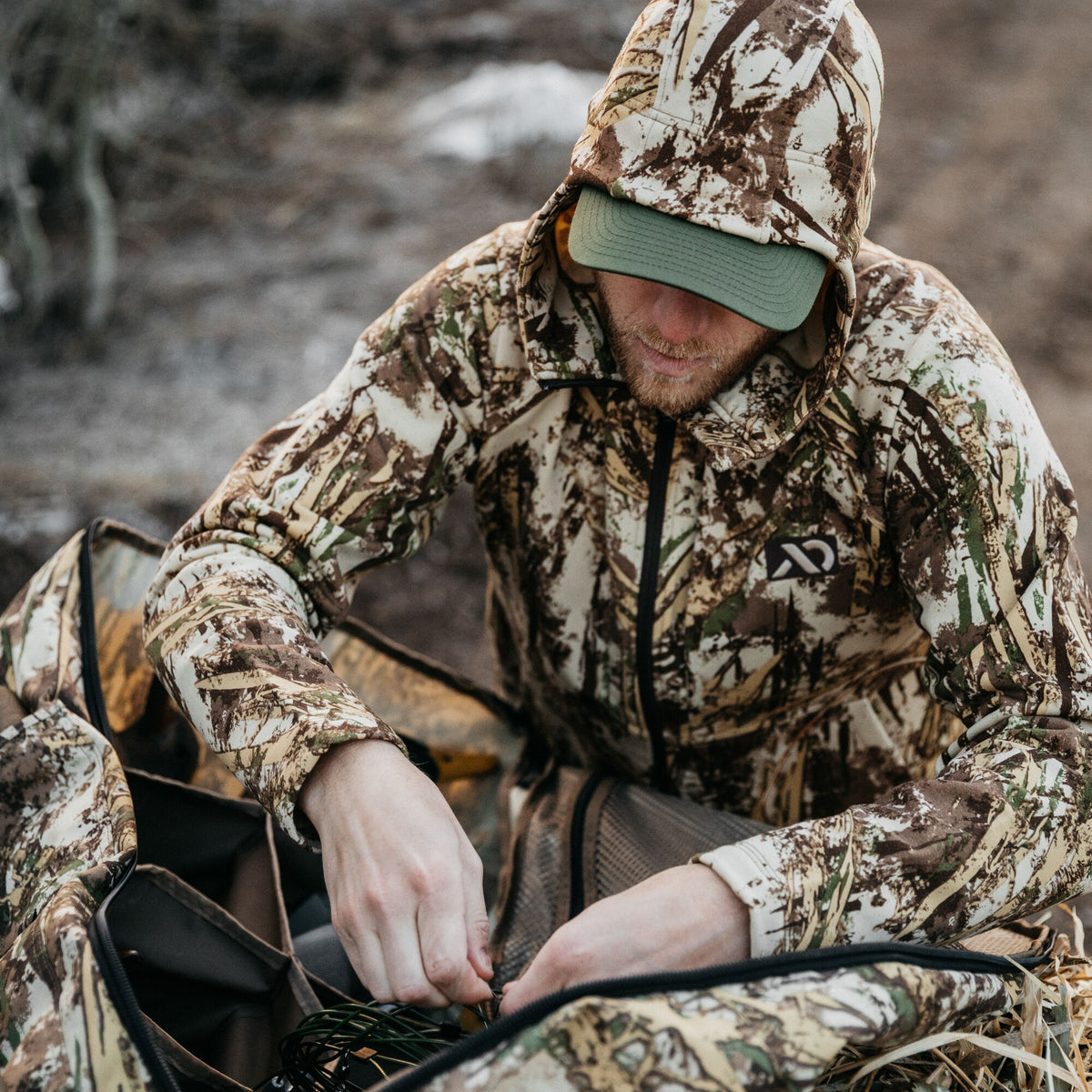 Man in a camouflage hooded jacket with a green baseball cap organizing items in a matching camouflage bag.