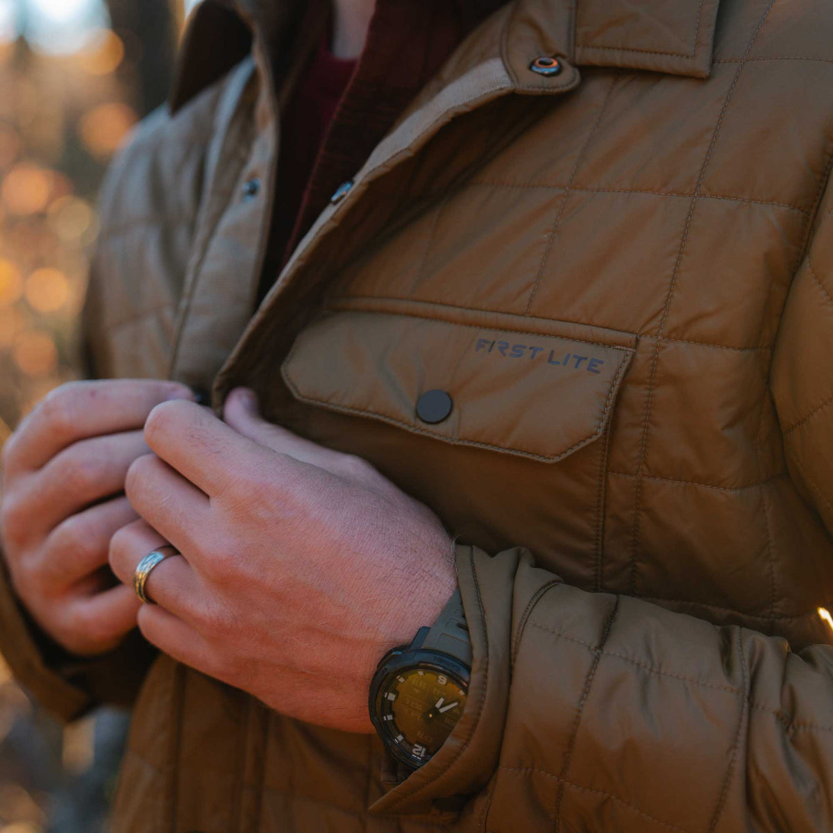 Close-up of a person buttoning a brown quilted jacket featuring a chest pocket with the "FIRST LITE" logo and snap buttons.