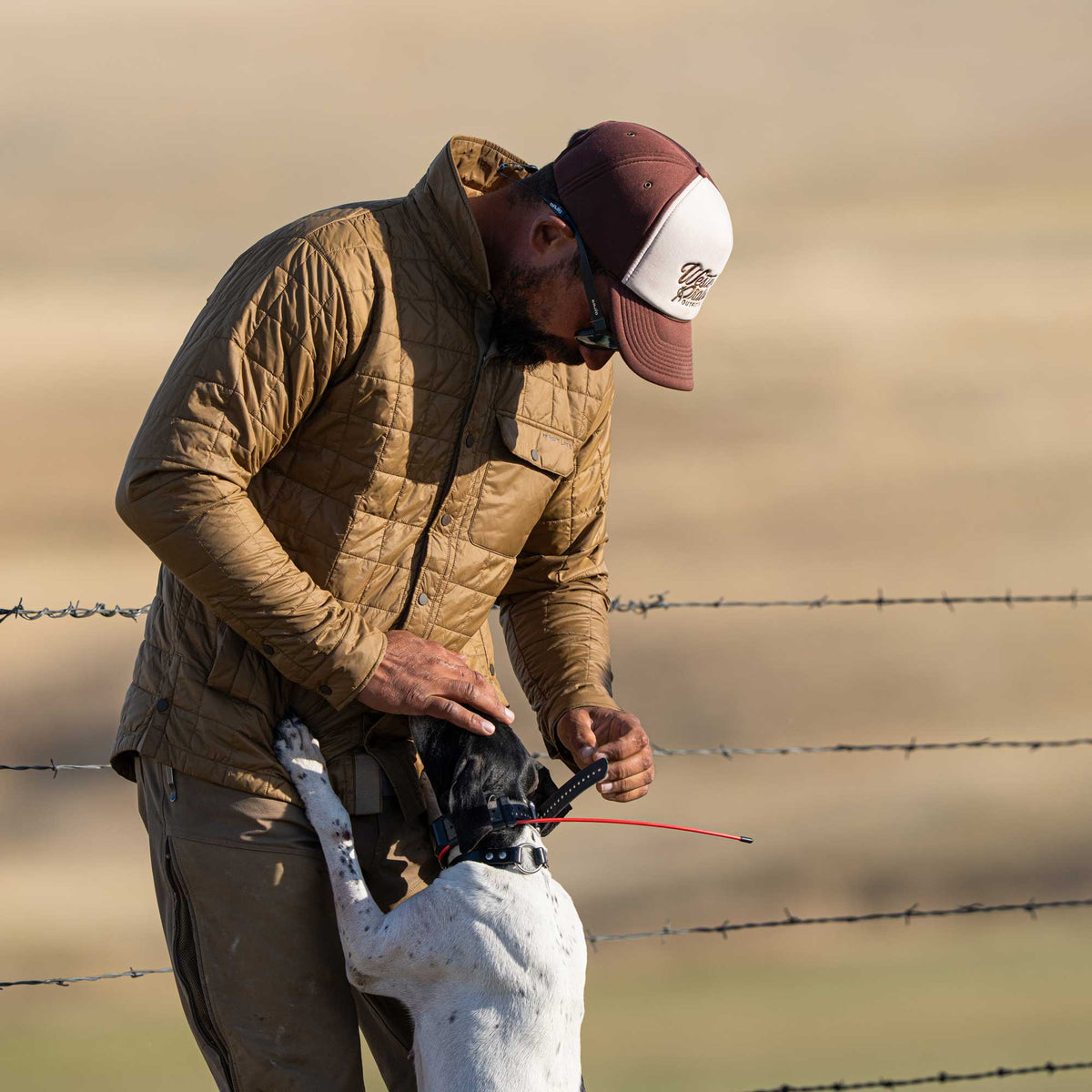 A man wears a tan quilted button-up jacket with a 'FIRST LITE' logo on its button-flap chest pocket, petting a dog.