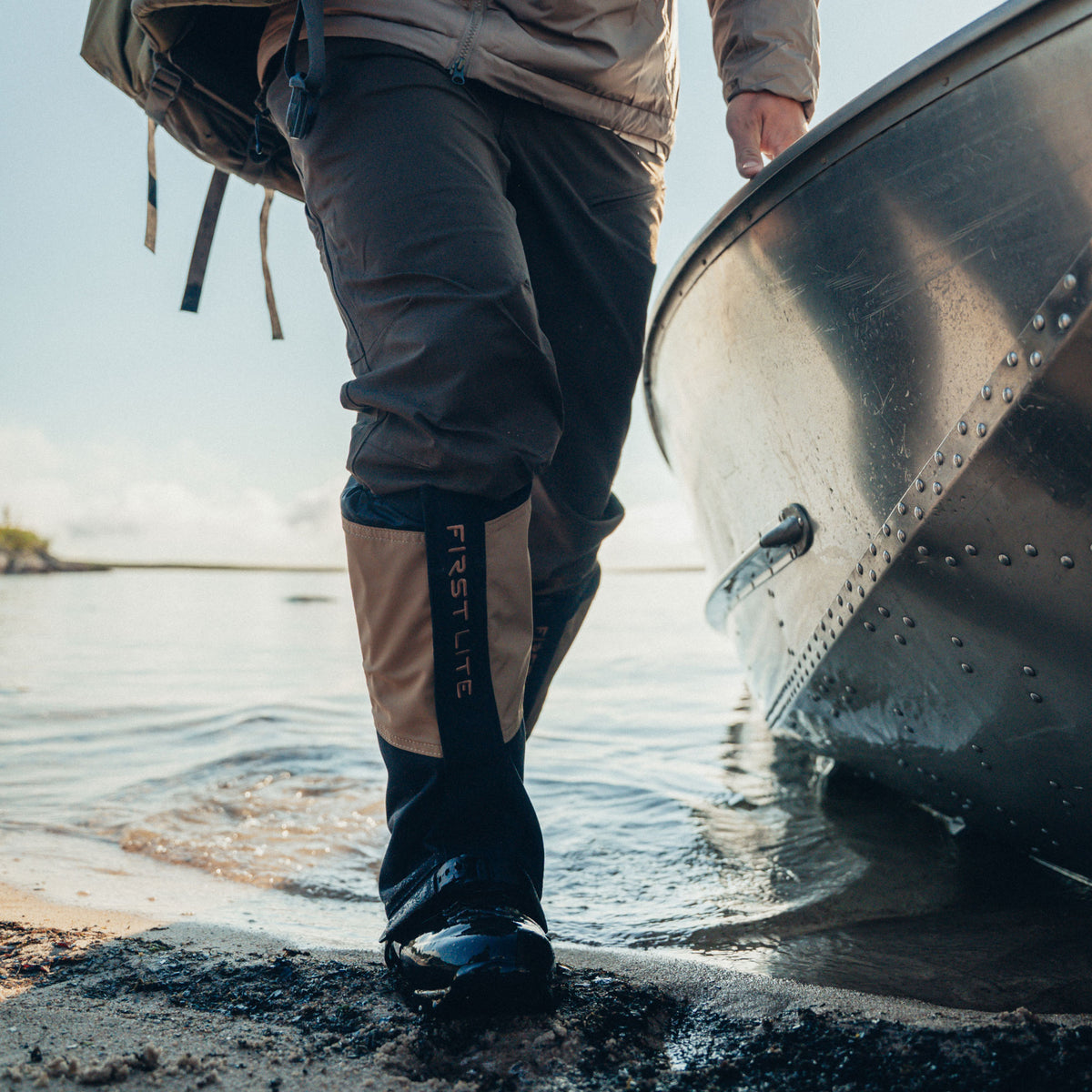 Man wearing boot gaiters, wading out of a lake dragging a boat