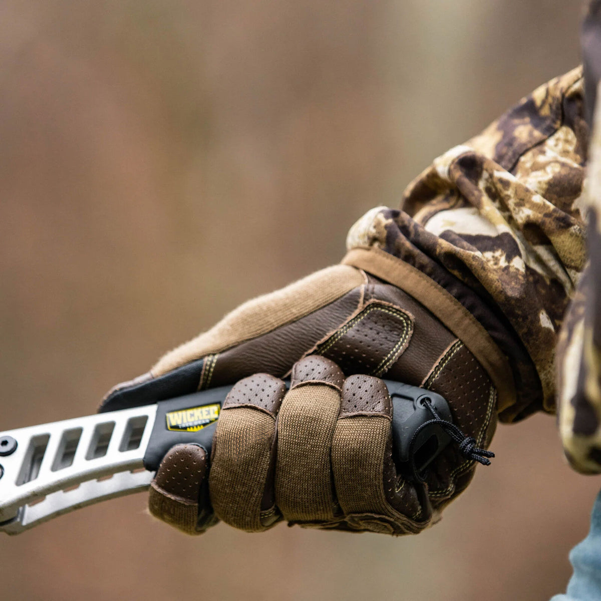 A hand in a brown and tan glove holds a black and silver Wicked Tree Gear pruning saw with a vented blade and yellow "WICKED" logo.