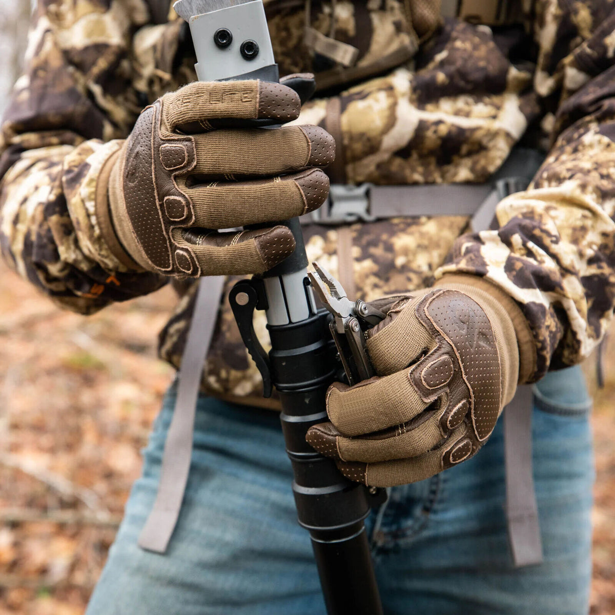 Two-toned brown and tan tactical gloves with textured knuckle and finger accents, worn by a person holding a pole and multi-tool.