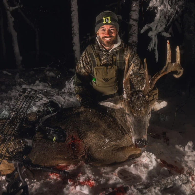 Smiling hunter in camo with large brown antlered deer and bow in snowy night.
