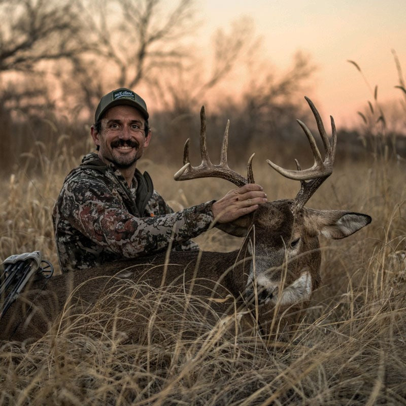 Smiling hunter in camo, green hat, with a large-antlered brown deer in tall grass.