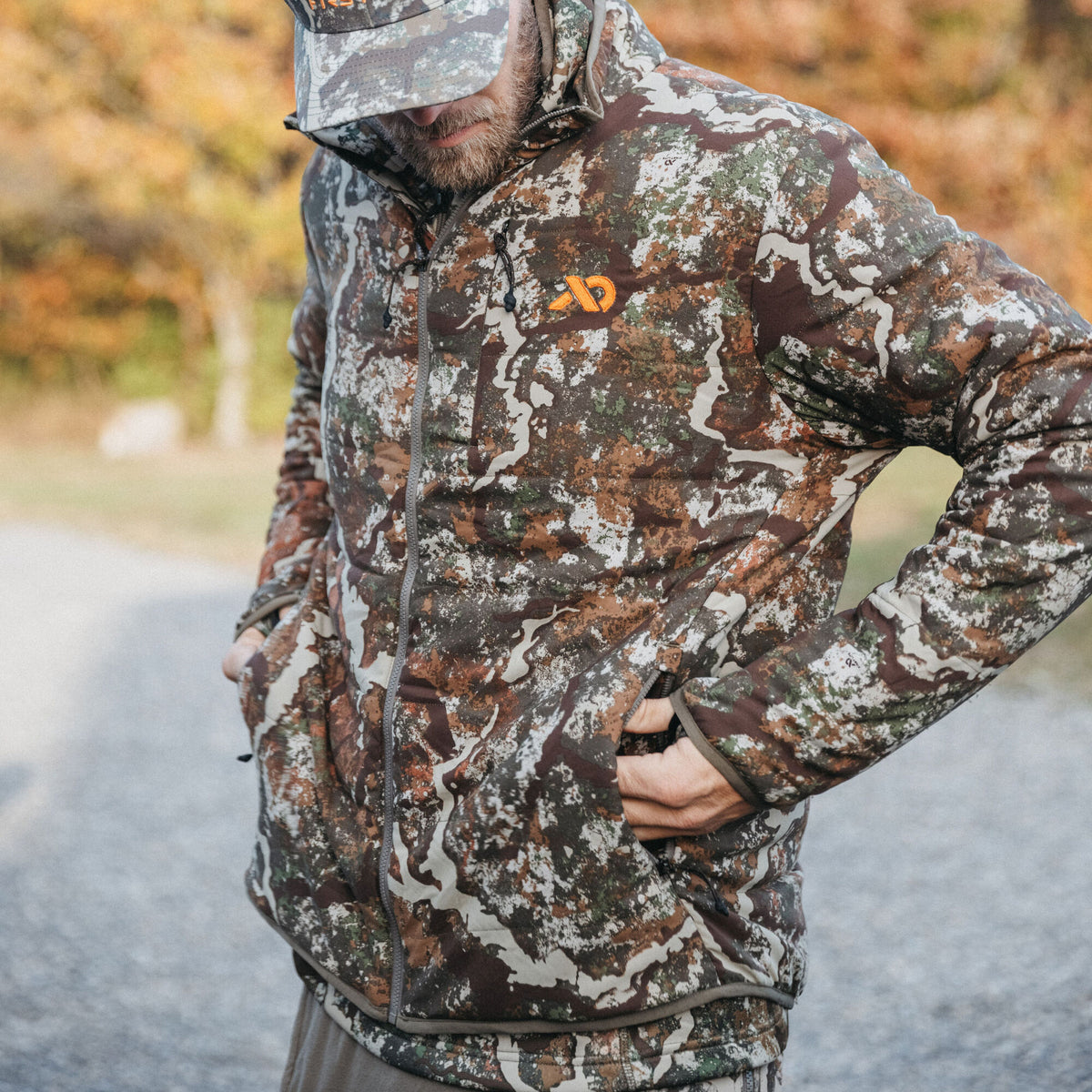 Full-zip insulated camouflage jacket with an orange chest logo and hands in front pockets.