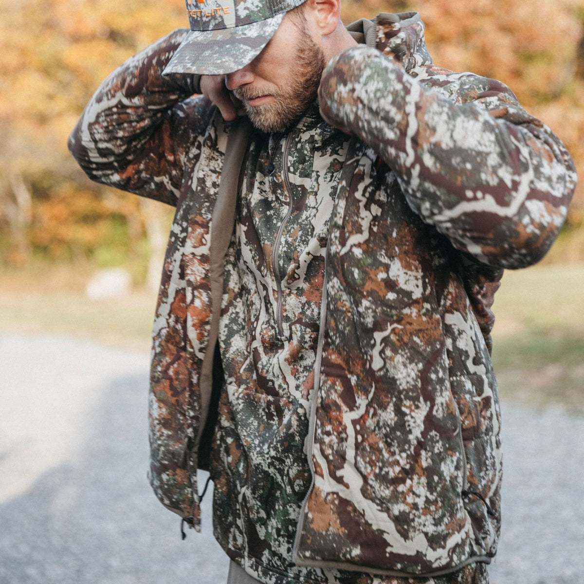 Man in a brown, green, and white camouflage full-zip jacket and matching cap, adjusting the jacket's collar.