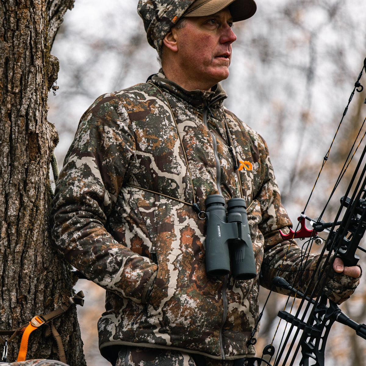 Man in a quilted multi-tone camouflage hunting jacket with an orange logo, holding a bow and binoculars, leaning on a tree.
