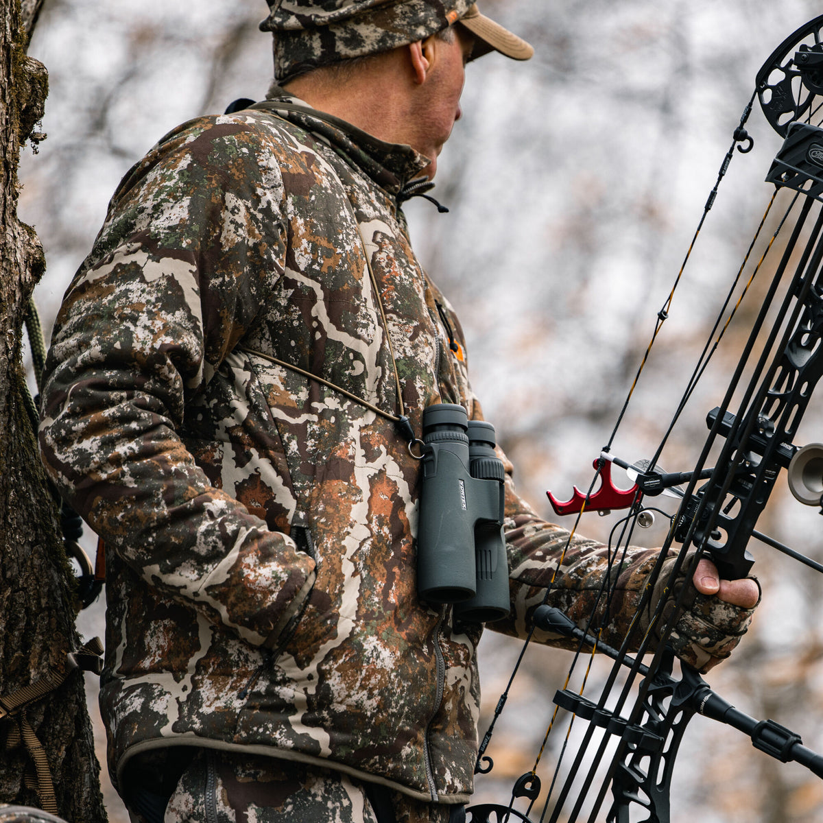 A hunter wears a brown, green, and white quilted camouflage jacket with a matching hat, holding a bow and wearing binoculars, in a tree stand.